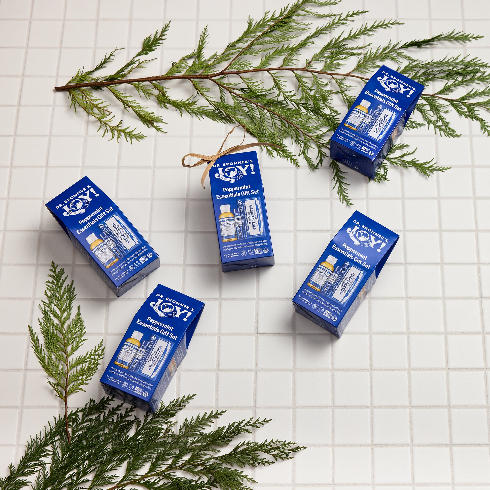 Dr. Bronners peppermint essential gift sets lay on a white tiled surface, surrounded by green pine branches. The packaging highlights Dr. Bronners eco friendly, vegan, organic soap products [One Size]