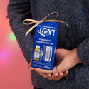 A person holds a blue gift box labeled Dr. Bronners Joy Peppermint Essentials Gift Set, tied with a ribbon, set against a festive, blurred pink background with lights [One Size]