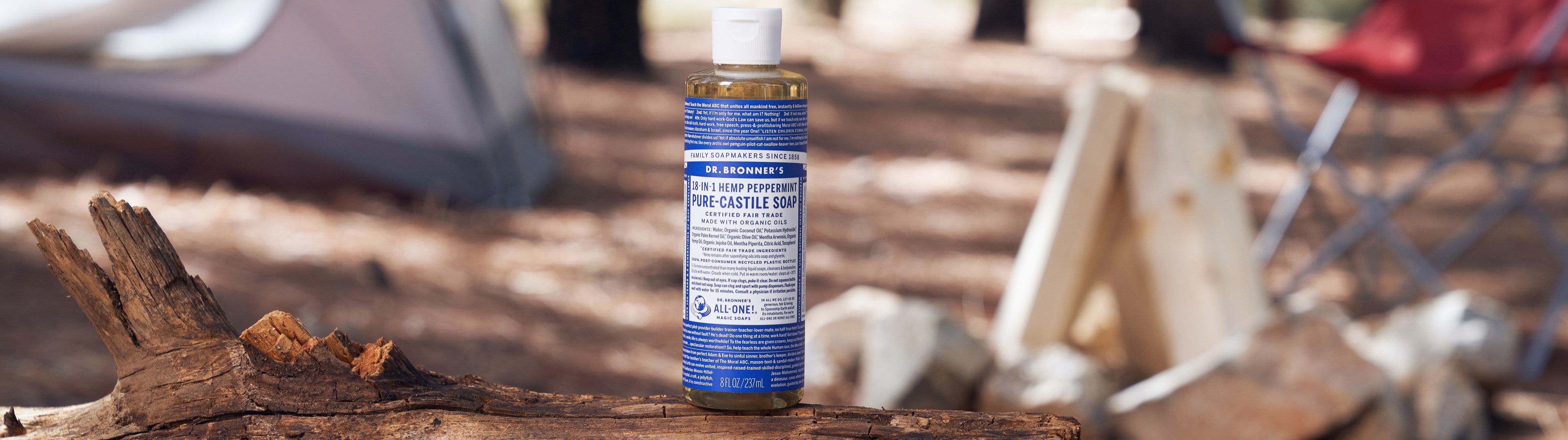 Dr. Bronners 18 in 1 Hemp Peppermint Pure Castile Soap bottle stands on a tree log. The backdrop features a camping setting with trees, a tent, and a red chair