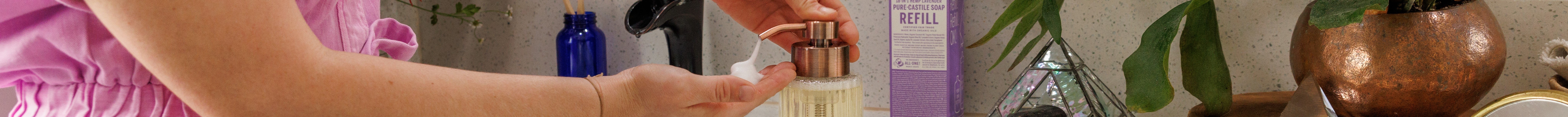 A person in a pink outfit pumps liquid soap into their hand from a glass dispenser. Nearby, a lavender Dr. Bronners Pure Castile Soap Refill box is on the counter with plants and a pyramid decoration
