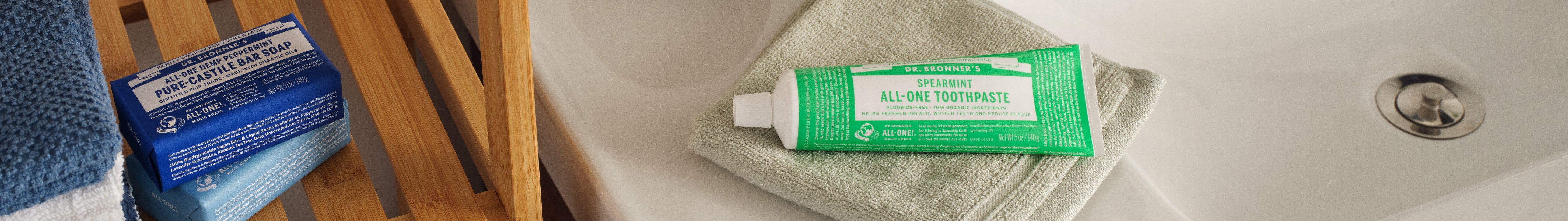 Dr. Bronners Spearmint All One Toothpaste sits on a green towel by a white sink. Nearby, a blue box of Dr. Bronners All One Hemp Peppermint Pure Castile Bar Soap is placed on wooden slats