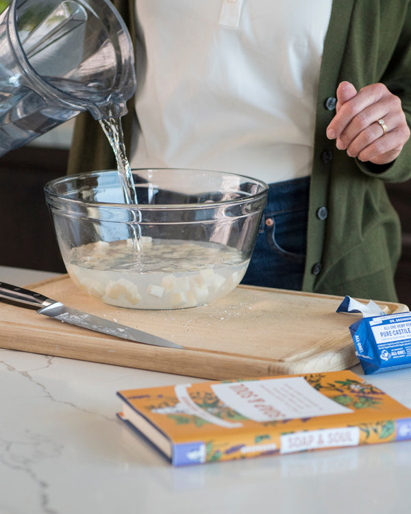 A person pours liquid into a clear bowl containing chopped ingredients on a kitchen counter. A Dr. Bronners Castile Soap box and a knife rest beside it. Vegan soap option displayed