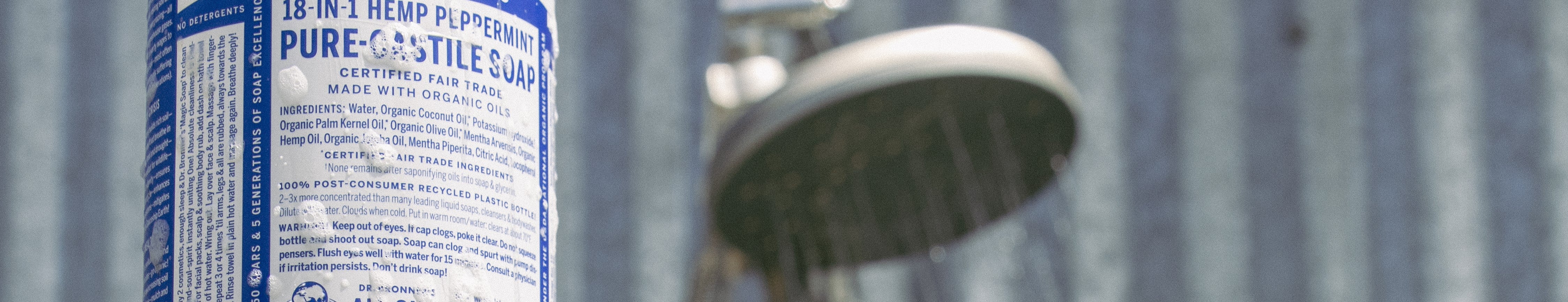 A bottle of Dr. Bronners 18 IN 1 Hemp Peppermint Pure Castile Soap stands upright near a showerhead text highlights certified fair trade and organic ingredients. Set against a metal wall backdrop
