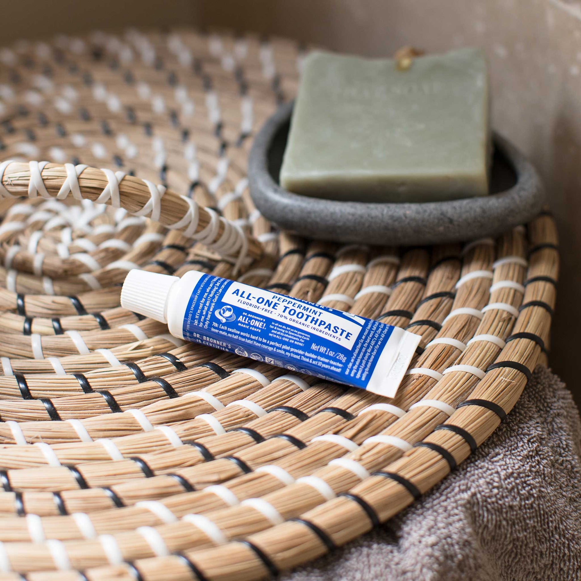 A small tube labeled Dr. Bronners Peppermint Organic Lip Balm lies on a woven mat next to a green bar of soap in a gray dish, creating a natural, eco friendly bathroom scene [5 oz]