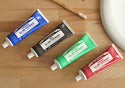 Four toothpaste tubes of various colors lie on a wooden surface next to a toothbrush and a sink. Text reads Dr. Bronners All One Toothpaste [5 oz]