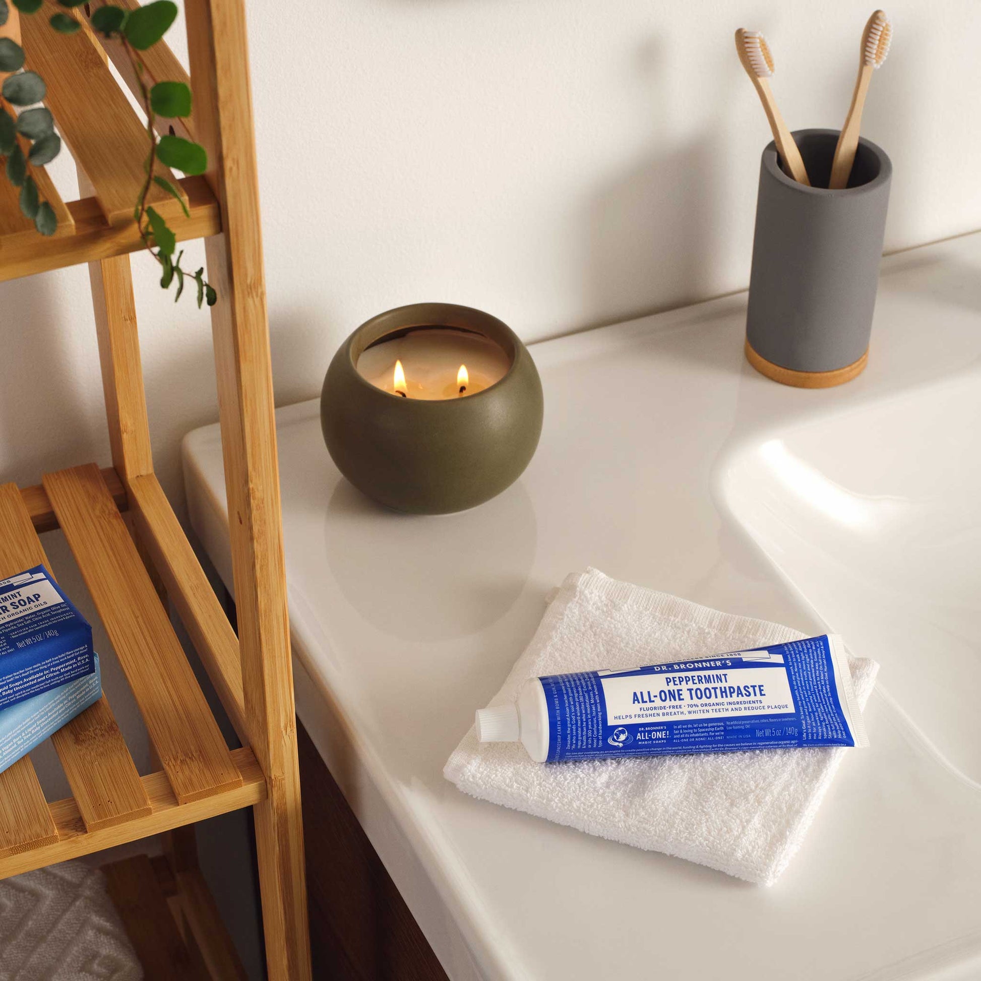 A package of Dr. Bronners unscented castile soap rests on a white towel by a bathroom sink. A lit candle and toothbrushes add ambiance, while shelves hold more products and plants [5 oz]