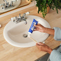 A person applies Dr Bronners toothpaste onto a toothbrush over a white sink with a wooden countertop. A potted plant and a polished faucet complete the tidy, eco friendly bathroom setting [5 oz]