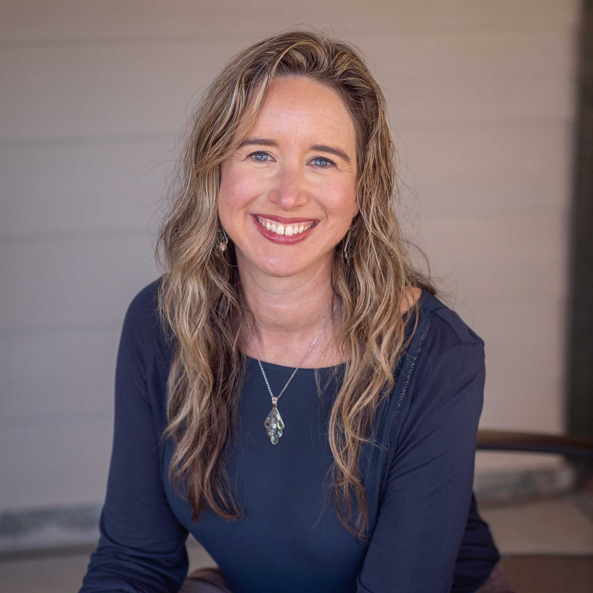 A person with long, wavy hair smiles warmly while sitting indoors. The background features a neutral toned wall, creating a welcoming atmosphere. No text present