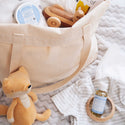 A canvas tote contains a wooden toy and baby items, including Dr. Bronners Baby Unscented Castile Soap. Nearby sit a plush fox toy, striped baby clothes, and pacifier on a patterned blanket