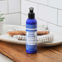 A blue bottle of Dr. Bronners Peppermint Organic Hair Crme sits on a wooden countertop. Nearby, a comb and folded towels create a bathroom setting, enhanced by a tiled wall backdrop [6 oz]