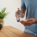 A person sprays Dr. Bronners Organic Peppermint Hand Cleansing Spray onto their palm, standing beside a wooden table adorned with a potted plant. DR. BRONNERS ORGANIC PEPPERMINT HAND CLEANSING SPRAY 60 ml