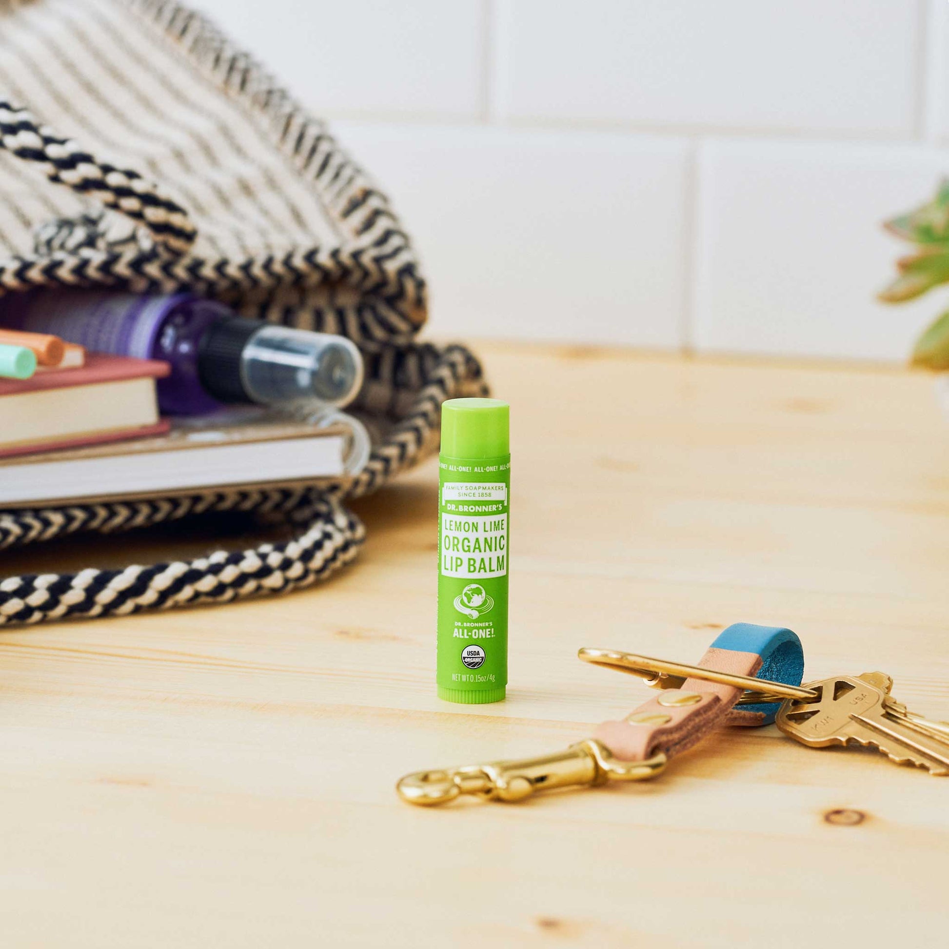 A lemon lime organic lip balm stands upright on a wooden surface beside a set of keys with colorful keychains. In the background, a patterned blanket and books create a cozy atmosphere [.15 oz]