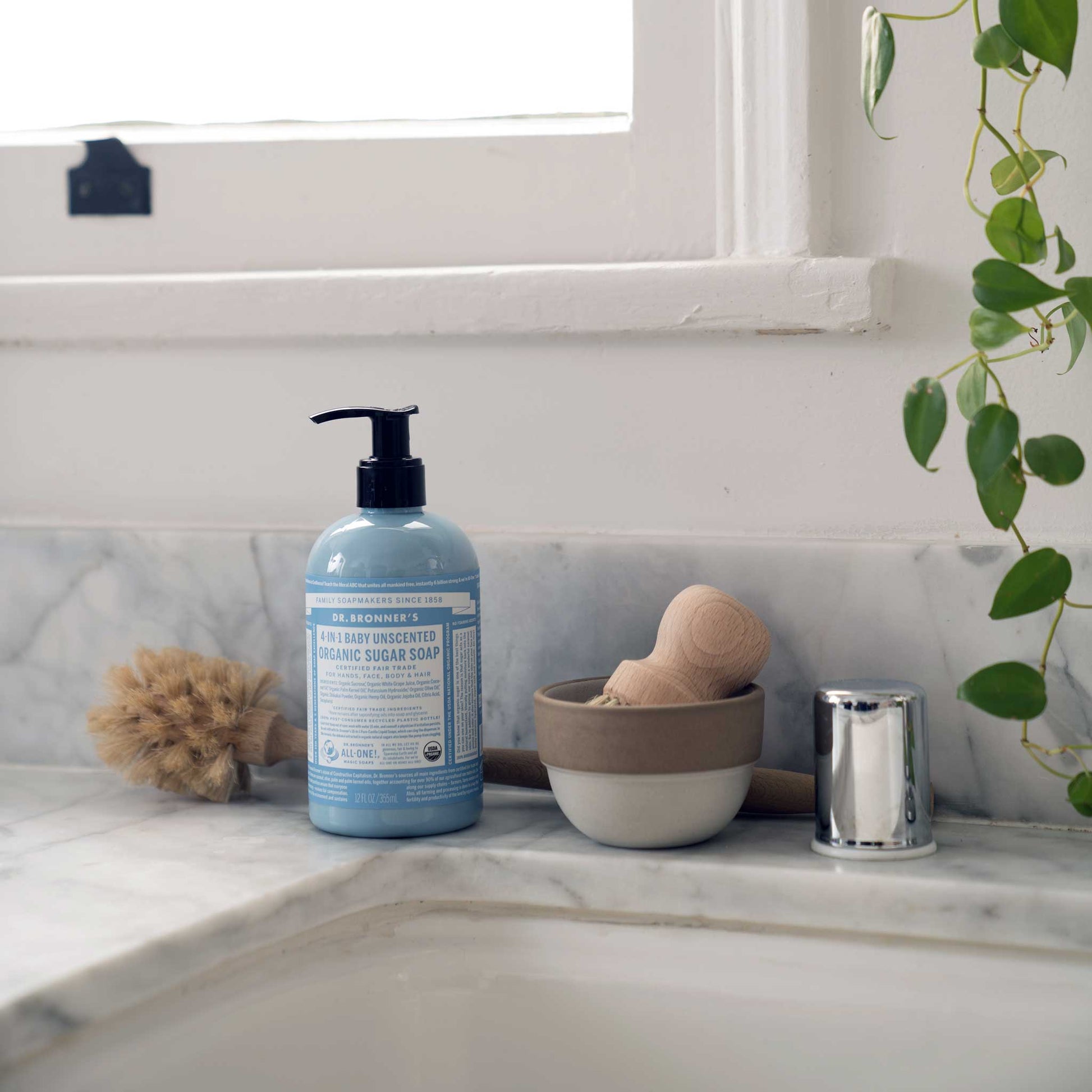 A bottle of Dr. Bronners 4 in 1 Baby Unscented Organic Sugar Soap stands on a marble bathroom counter beside a scrubbing brush, plant, and bowl. DR. BRONNERS 4 IN 1 BABY UNSCENTED ORGANIC SUGAR SOAP [12 oz]