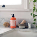 A bottle of Dr. Bronners citrus castile soap rests on a marble countertop beside a small bowl and a brush. Its next to a sink faucet, with a potted plant hanging nearby [12 oz]