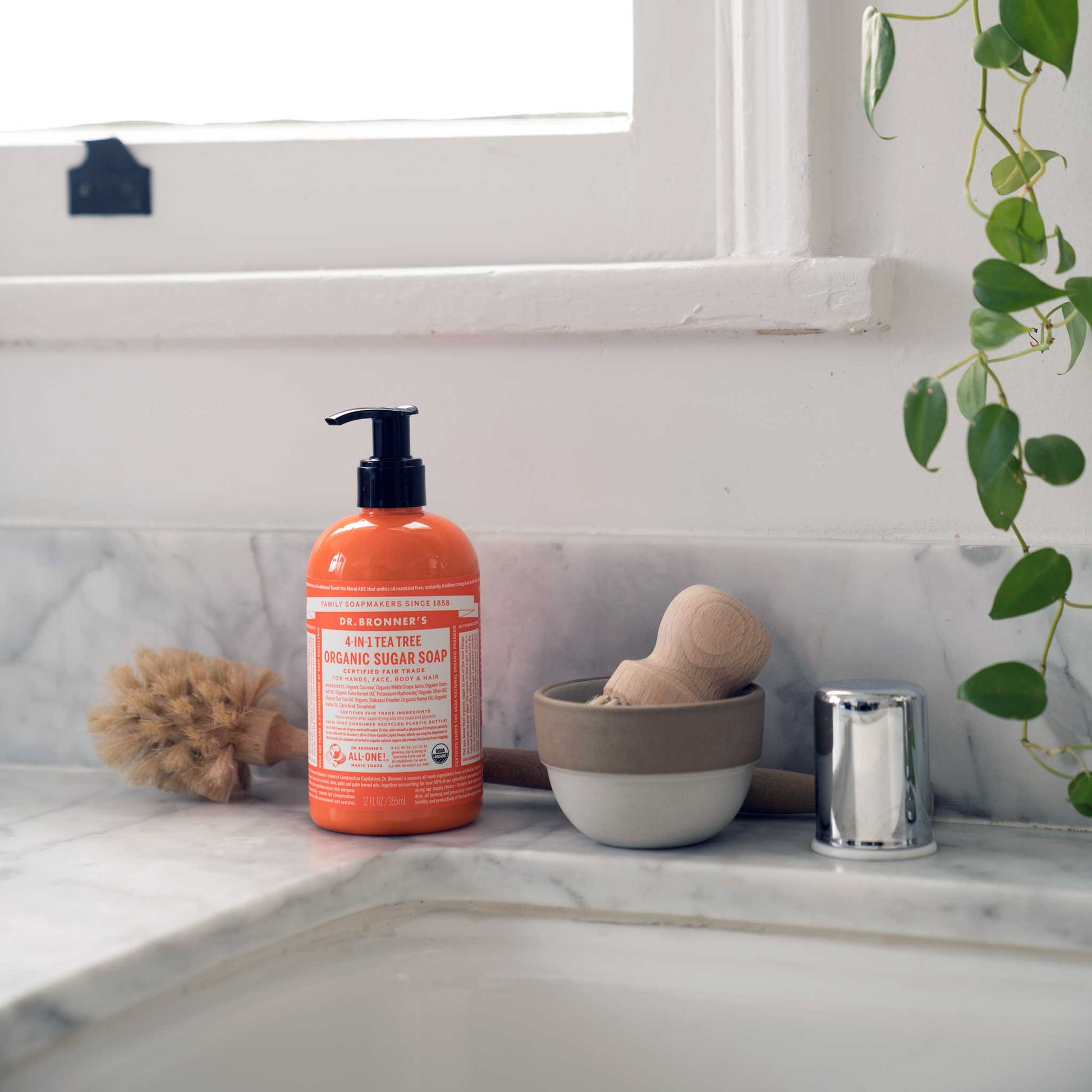 A bottle of Dr. Bronners citrus castile soap rests on a marble countertop beside a small bowl and a brush. Its next to a sink faucet, with a potted plant hanging nearby [12 oz]