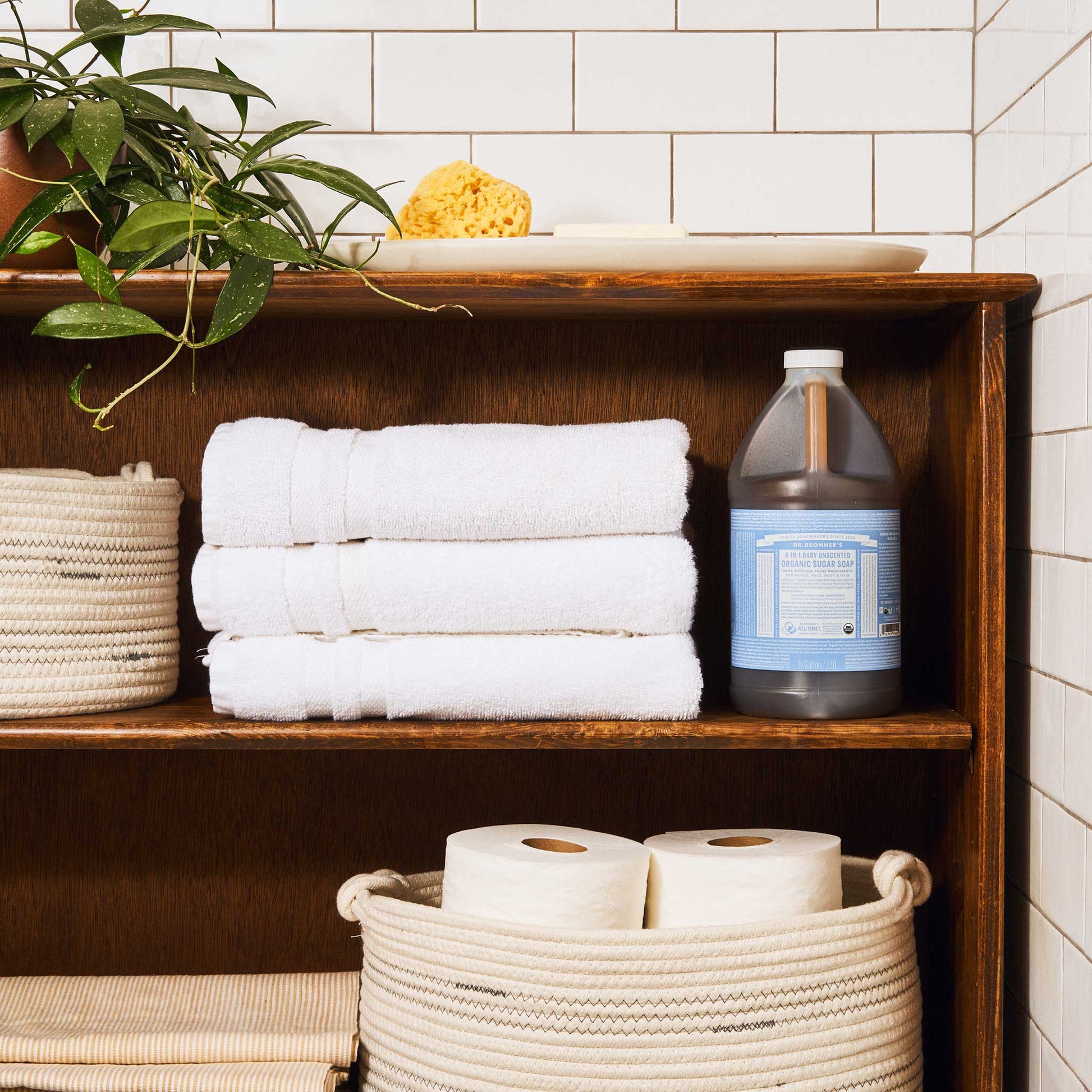 A bottle of Dr. Bronners liquid castile soap with a blue label sits on a wooden shelf. Surrounding it are neatly stacked white towels, a potted plant, and white tiles [1/2 Gallon]