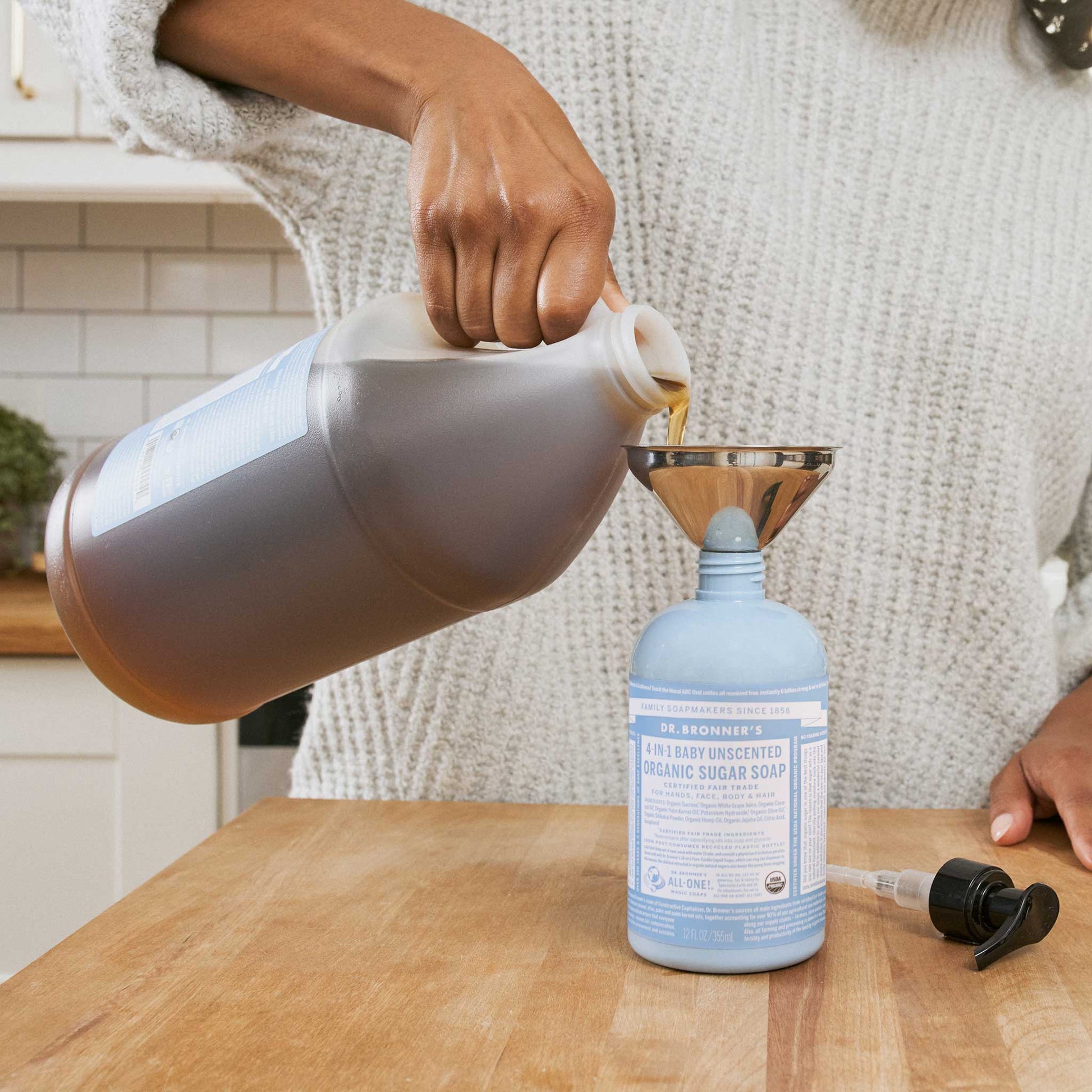 A person pours Dr Bronners 18 in 1 Baby Unscented Organic Sugar Soap from a large jug through a funnel into a smaller bottle on a wooden kitchen counter [12 oz]