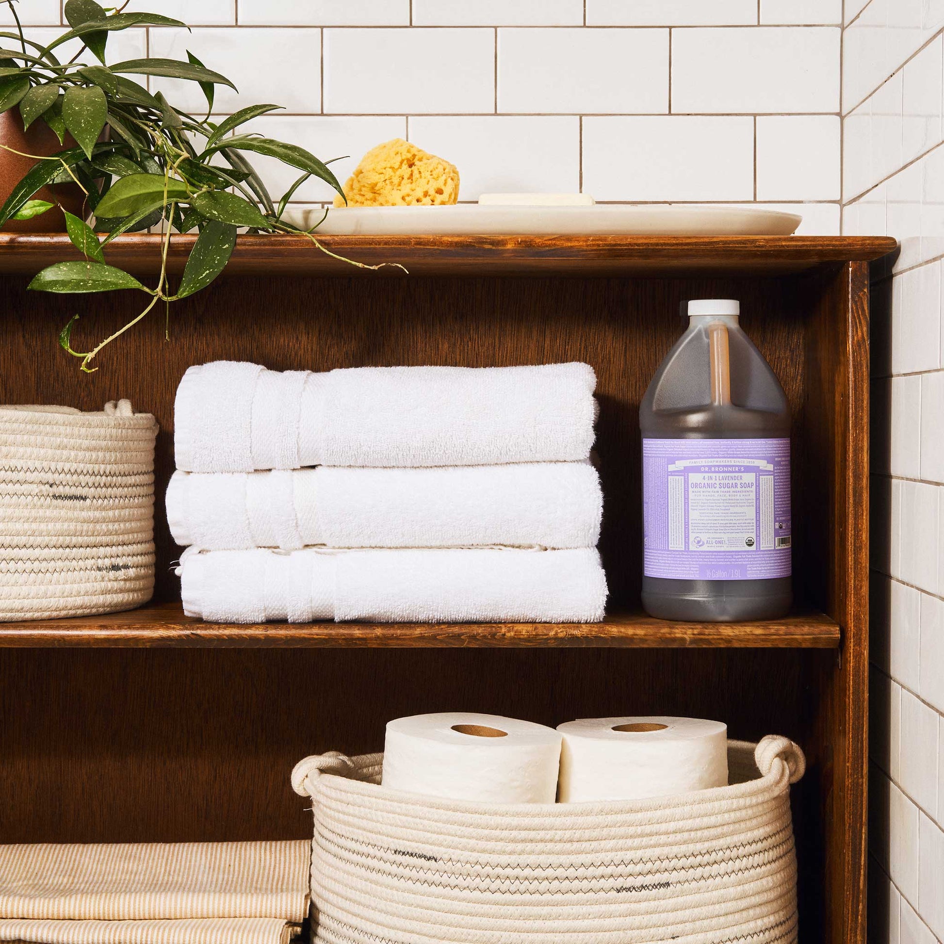 A wooden shelf in a tiled bathroom holds neatly stacked white towels, a bottle of Dr. Bronners lavender castile soap, toilet paper in baskets, a plant, and a sponge on top [1/2 Gallon]