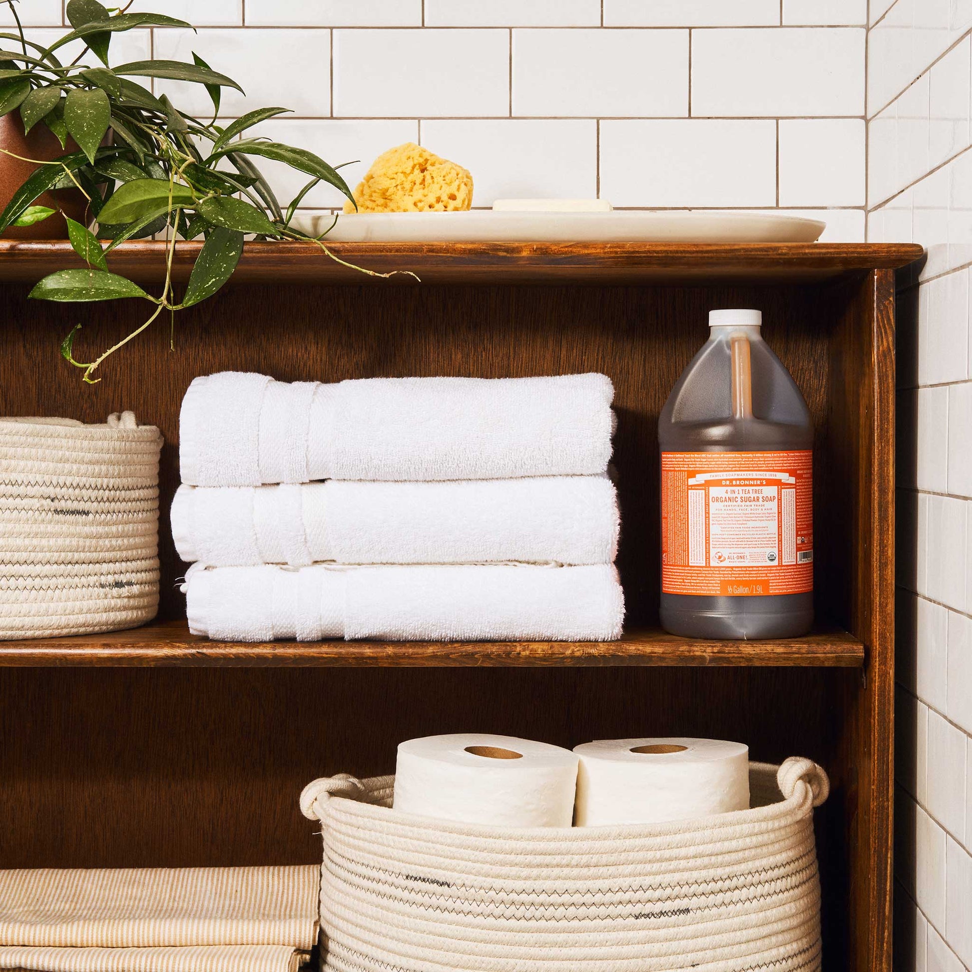 A wooden bathroom shelf displays a large bottle of Dr. Bronners organic sugar soap, white towels, and toilet paper, surrounded by a plant and tile walls, creating a tidy, natural setting [1/2 Gallon]