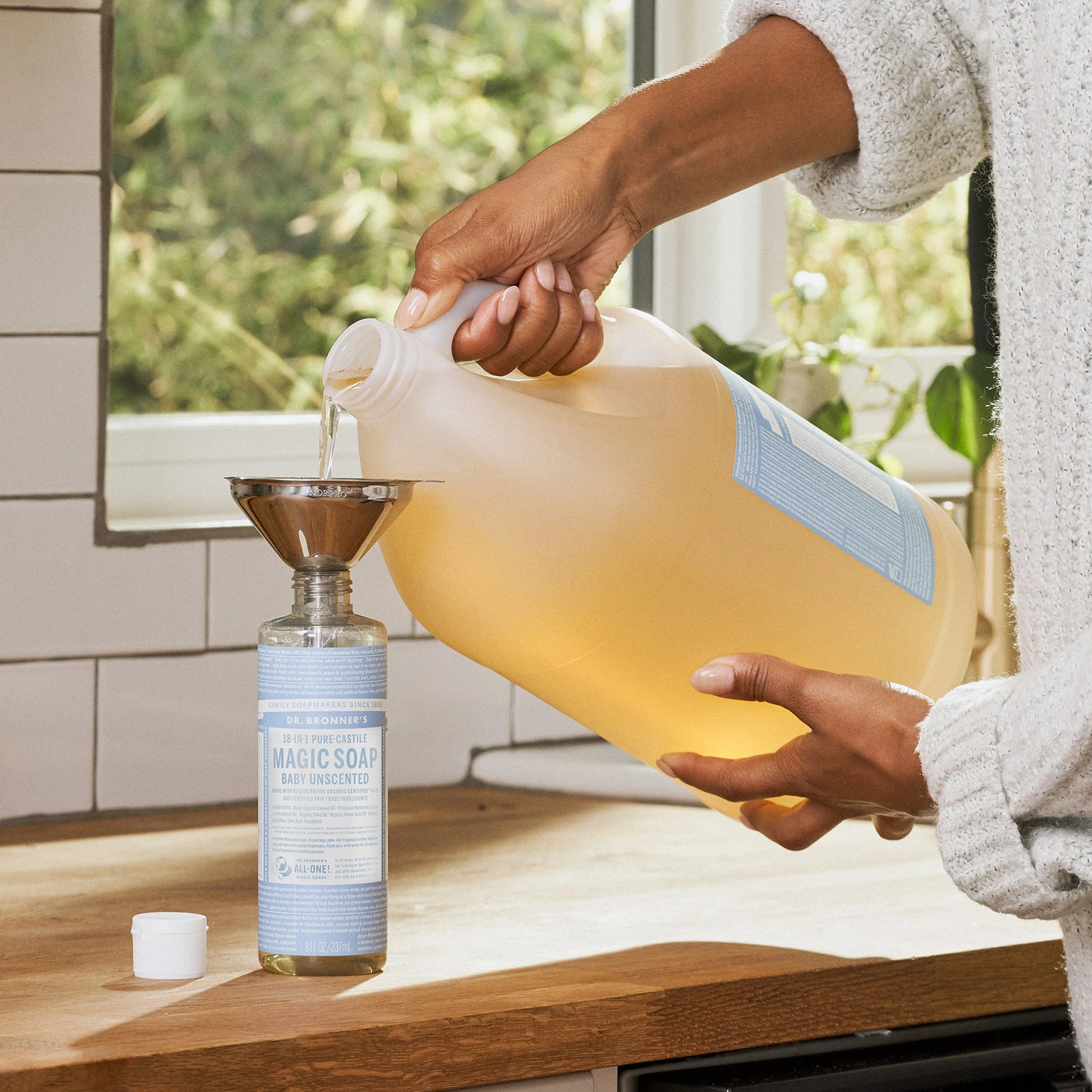 A person pours Dr. Bronners Baby Unscented Pure Castile Liquid Soap from a large bottle into a smaller bottle using a funnel on a kitchen counter, creating an eco friendly refill setting [16 oz, 1 Gallon]