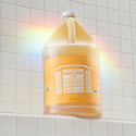 A large bottle of Dr. Bronners 18 in 1 Citrus Magic Soap sits on a tiled surface, illuminated by a rainbow light. The label details various uses, emphasizing eco friendly and organic ingredients, and the bottle capacity reads 1 Gallon 3.8 L [1 Gallon]