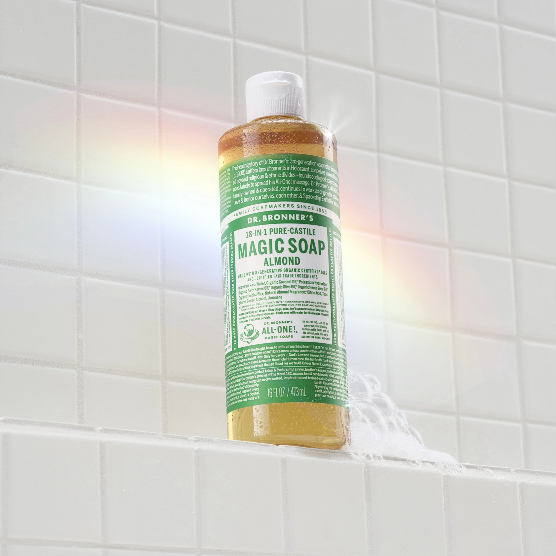 A bottle of Dr. Bronners eucalyptus castile soap stands on a white tiled shower ledge, with a soapy trail beneath it. A rainbow reflection appears on the tiles [16 oz]