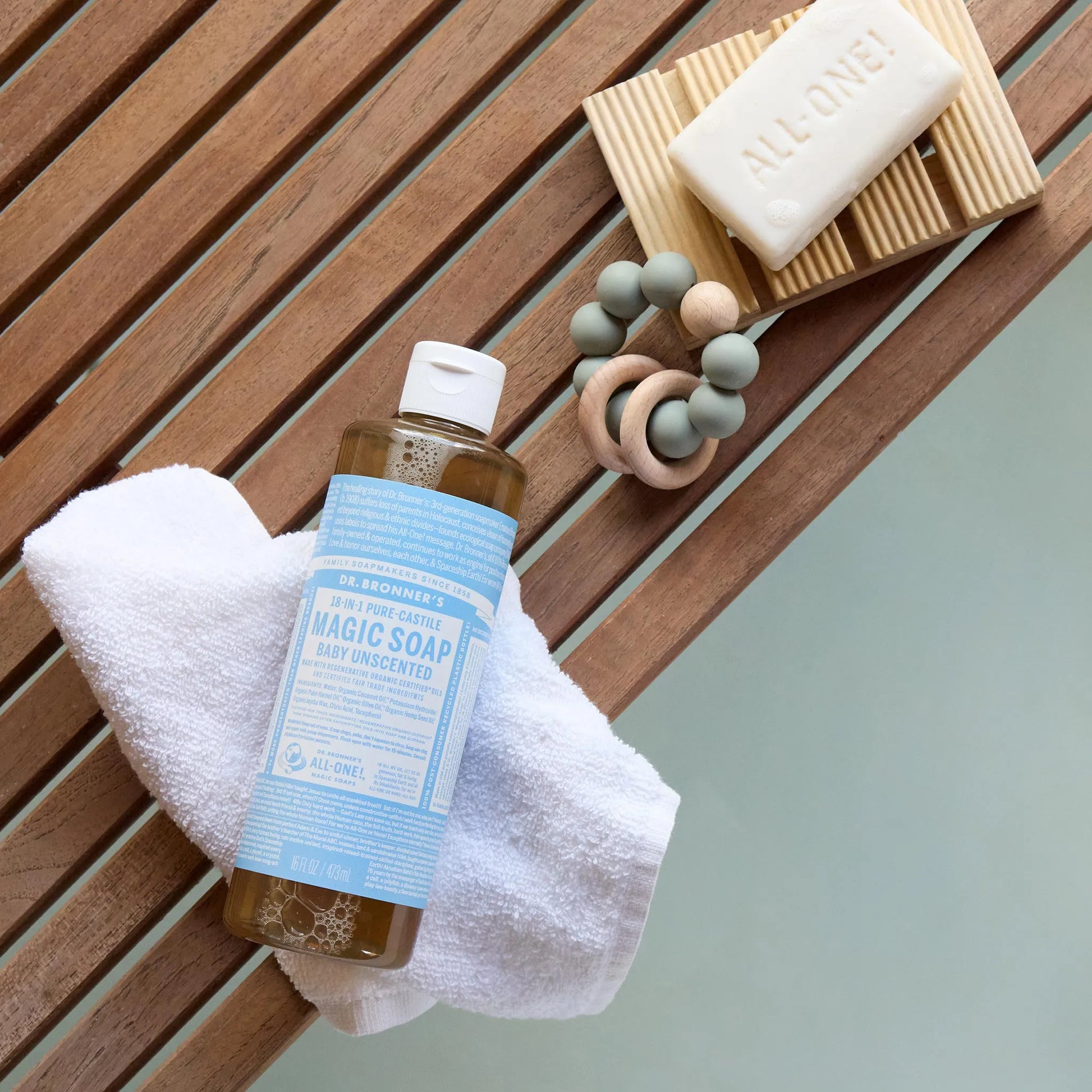 A bottle of Dr. Bronners Baby Unscented Pure Castile Soap rests on a white towel on a wooden surface, accompanied by a bar of soap and a beaded ring [16 oz]