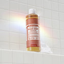A bottle of Dr. Bronners 18 in 1 Pure Castile Magic Soap in eucalyptus scent rests on a white tiled bathroom shelf, surrounded by colorful light reflections and suds. Text reads DR. BRONNERS 18 IN 1 PURE CASTILE MAGIC SOAP EUCALYPTUS. Made with regenerative organic certified oils and certified fair trade ingredients for the betterment of people and planet [16 oz]