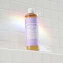 A bottle of Dr. Bronners lavender liquid castile soap sits on a tiled shower shelf, illuminated by a soft rainbow light. The label features text highlighting its organic, eco friendly ingredients [16 oz]