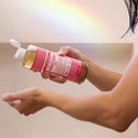 A person dispenses liquid from a pink bottle of Dr. Bronners pure castile soap into their palm, under a soft rainbow light in a bathroom setting [16 oz]