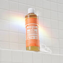 A bottle of Dr. Bronners Citrus Pure Castile Liquid Soap stands on a white tiled shelf, illuminated by a rainbow reflection. Ideal for eco friendly, vegan personal care routines [16 oz]