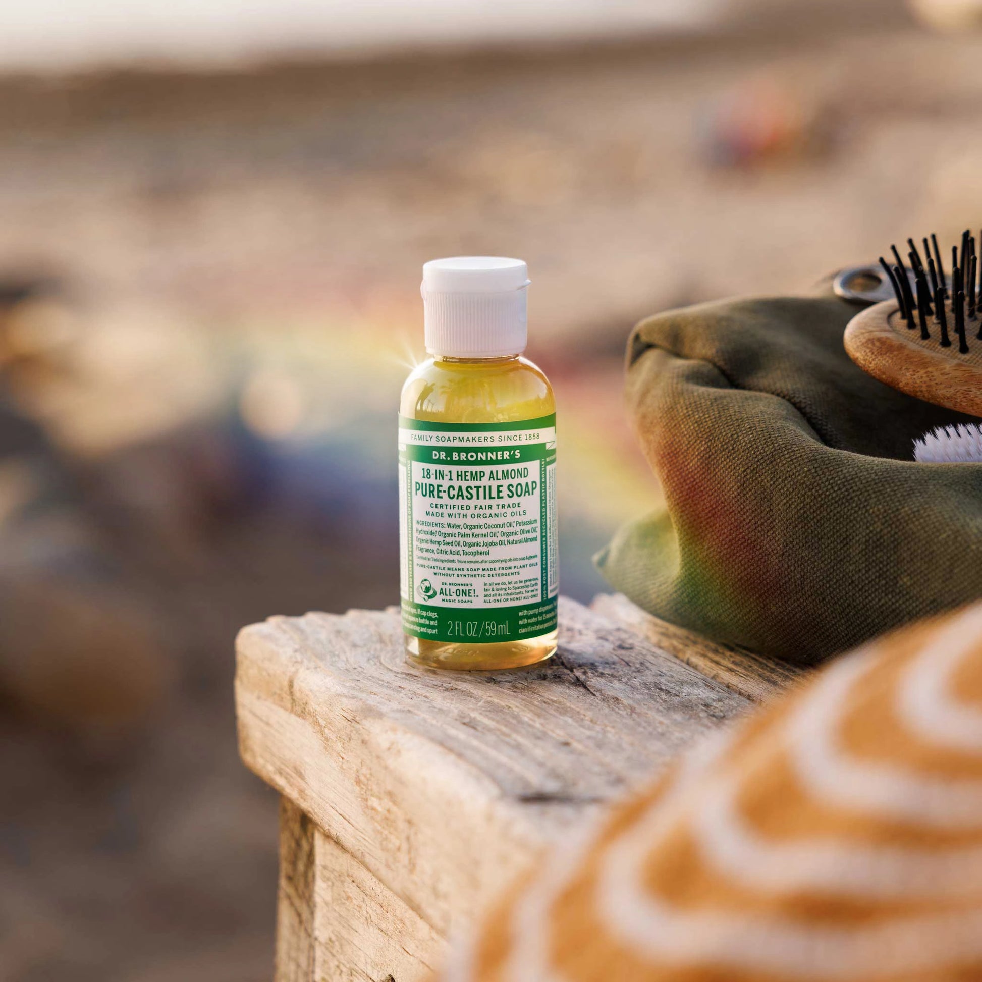 A small bottle of Dr. Bronners 18 in 1 Hemp Almond Pure Castile Soap rests on a rustic wooden surface near a beach. Text includes Certified Fair Trade and Made with Organic Oils, 2 fl oz [2 oz]