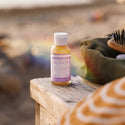 A small bottle of Dr. Bronners 18 in 1 Hemp Lavender Pure Castile Soap rests on a wooden surface at a beach. Nearby, a colorful bag and a brush add context [2 oz]