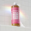 A bottle of Dr. Bronners rose castile soap stands on a white tiled shelf, reflecting sunlight to create a rainbow effect. The setting suggests a bright, clean bathroom [32 oz]