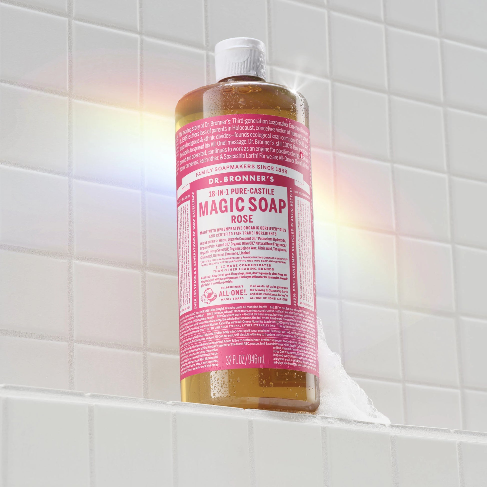 A bottle of Dr. Bronners rose castile soap stands on a white tiled shelf, reflecting sunlight to create a rainbow effect. The setting suggests a bright, clean bathroom [32 oz]