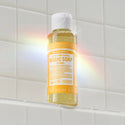 A bottle of Dr. Bronners Citrus Pure Castile Magic Soap sits on a white tiled shelf, illuminated by a rainbow light. Text reads 18 IN 1 Pure Castile MAGIC SOAP Citrus 3 FL OZ 89 mL [3.4 oz]