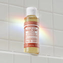 A bottle of Dr. Bronners 18 in 1 Pure Castile Magic Soap Eucalyptus sits on a white tiled shelf, with a rainbow reflection on the surface. Text includes DR. BRONNERS, 18 IN 1 PURE CASTILE, MAGIC SOAP, EUCALYPTUS, ALL ONE, 3.4 FL OZ 100 ml [3.4 oz]