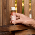 A hand holds a small bottle of Dr. Bronners 18 in 1 Pure Castile Magic Soap against a wooden fence. The label features product details in a natural outdoor setting [3.4 oz]