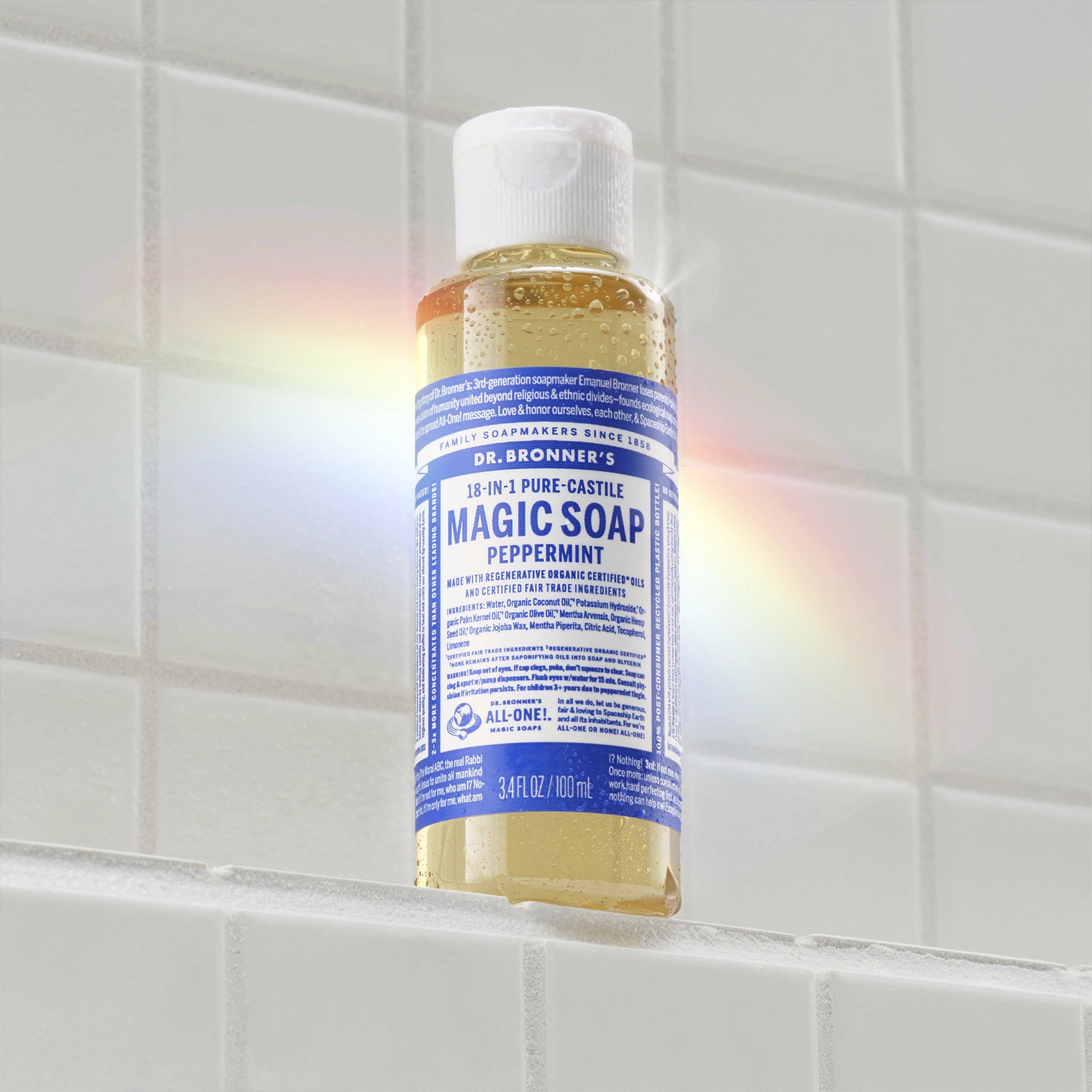A bottle of Dr. Bronners 18 in 1 Pure Castile Magic Soap Peppermint is standing upright on a tiled shelf. A subtle rainbow reflection appears on the bottle, highlighting its eco friendly design [3.4 oz]