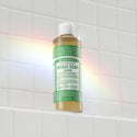 A bottle of Dr. Bronners 18 in 1 Pure Castile Magic Soap stands on a white tiled shelf, illuminated by a rainbow light. Text includes product information and usage instructions [8 oz]