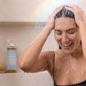 A person enjoys a shower, massaging their head under streaming water with Dr. Bronners Magic Soap nearby on a shelf in a minimalistic bathroom [32 oz]