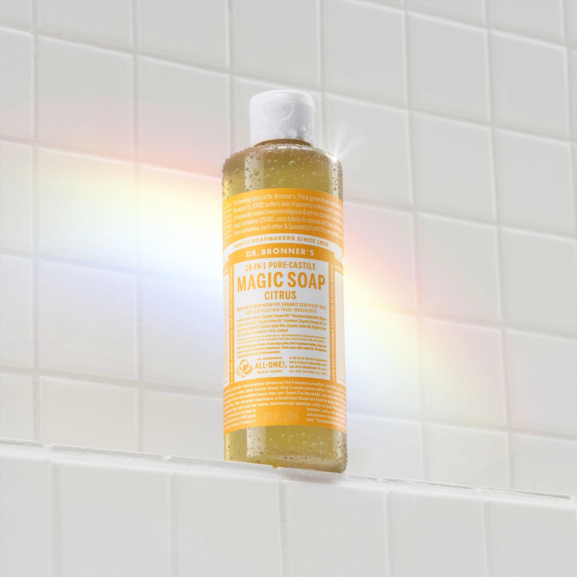 A yellow bottle of Dr. Bronners citrus castile soap sits against a tiled wall, illuminated by a rainbow light reflection. The label features white text detailing the soaps natural ingredients and eco friendly qualities [8 oz]