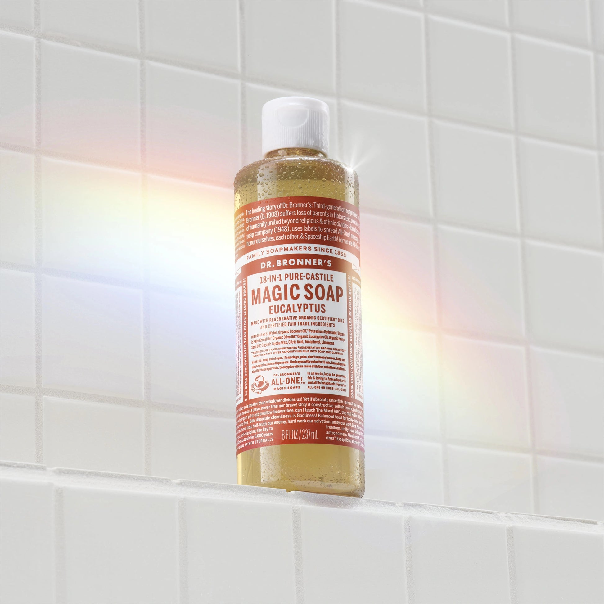 A bottle of Dr. Bronners Castile Soap with a red label rests against white tiled wall, casting a soft rainbow reflection. Multipurpose, eco friendly body wash and cleaner [8 oz]