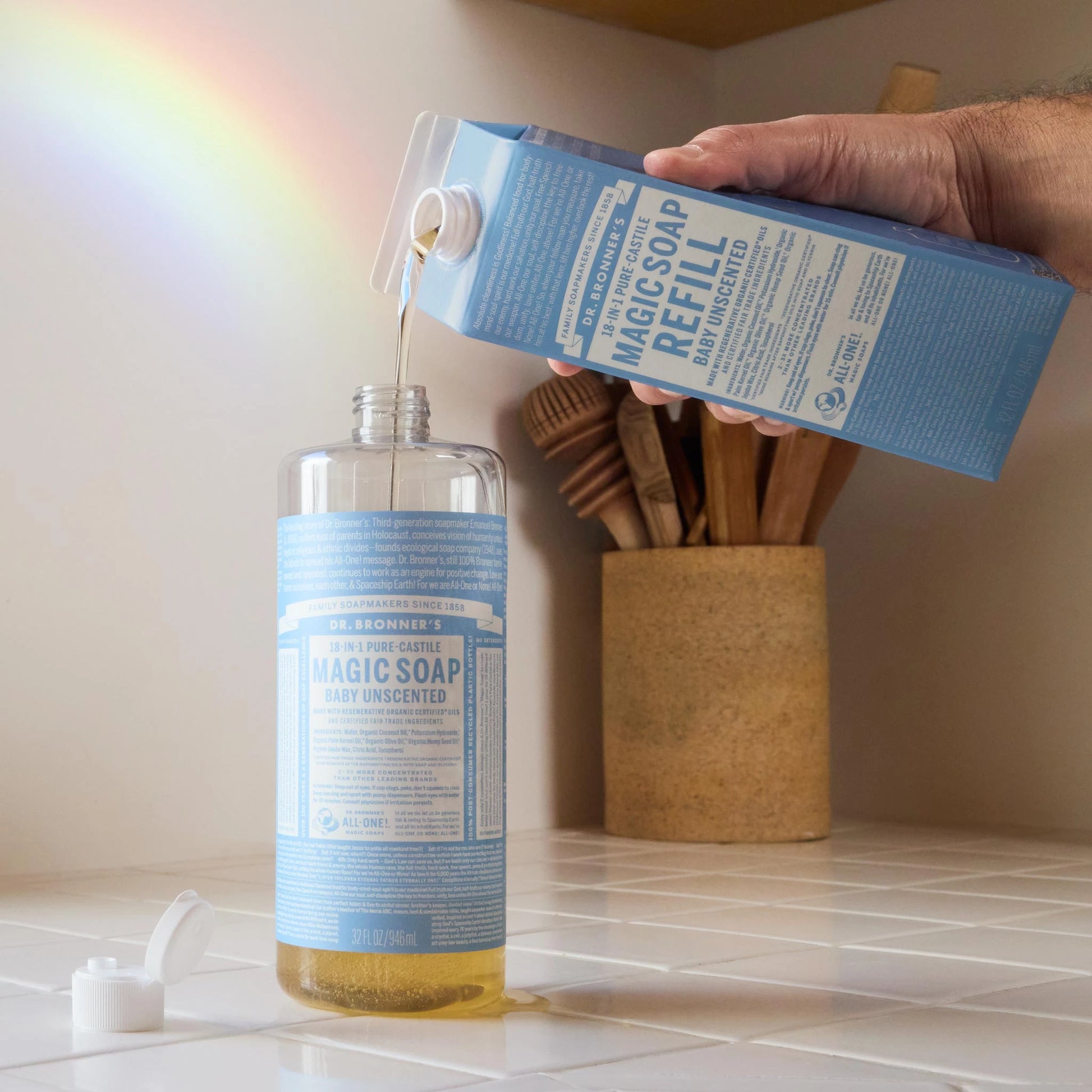 A bottle of Dr. Bronners baby unscented castile soap being refilled on a kitchen counter, with a rainbow reflection on the wall. A container with utensils is in the background [32 oz]