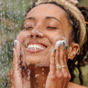 A person gently lathers their face with soap under a shower, smiling blissfully. Bubbles cover their cheeks, set against a blurred green outdoor background, suggesting a refreshing experience with natural castile soap  [2 oz, 3.4 oz, 4 oz, 8 oz, 16 oz, 32 oz, 1/2 Gallon, 1 Gallon]