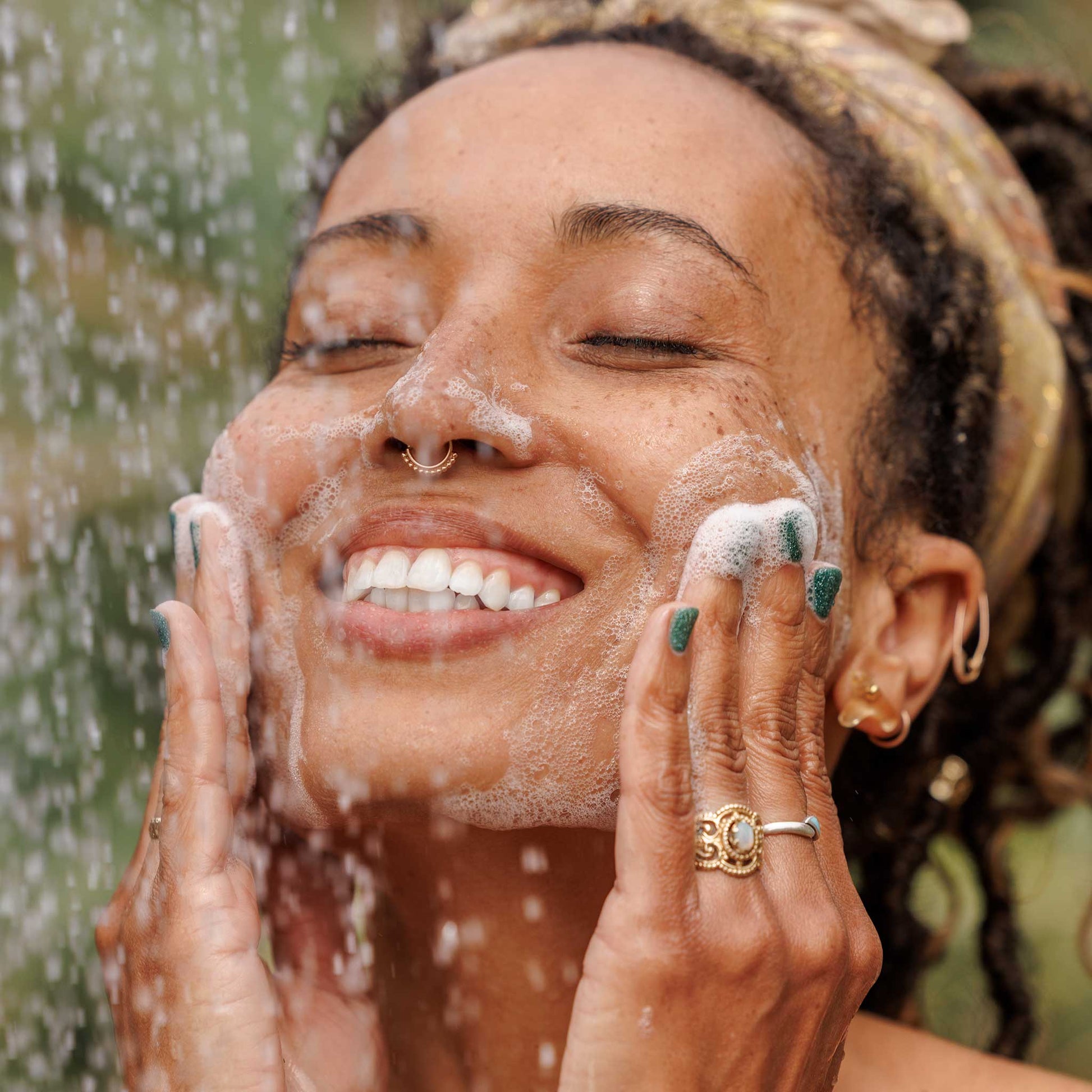 A person gently lathers their face with soap under a shower, smiling blissfully. Bubbles cover their cheeks, set against a blurred green outdoor background, suggesting a refreshing experience with natural castile soap  [2 oz, 3.4 oz, 4 oz, 8 oz, 16 oz, 32 oz, 1/2 Gallon, 1 Gallon]
