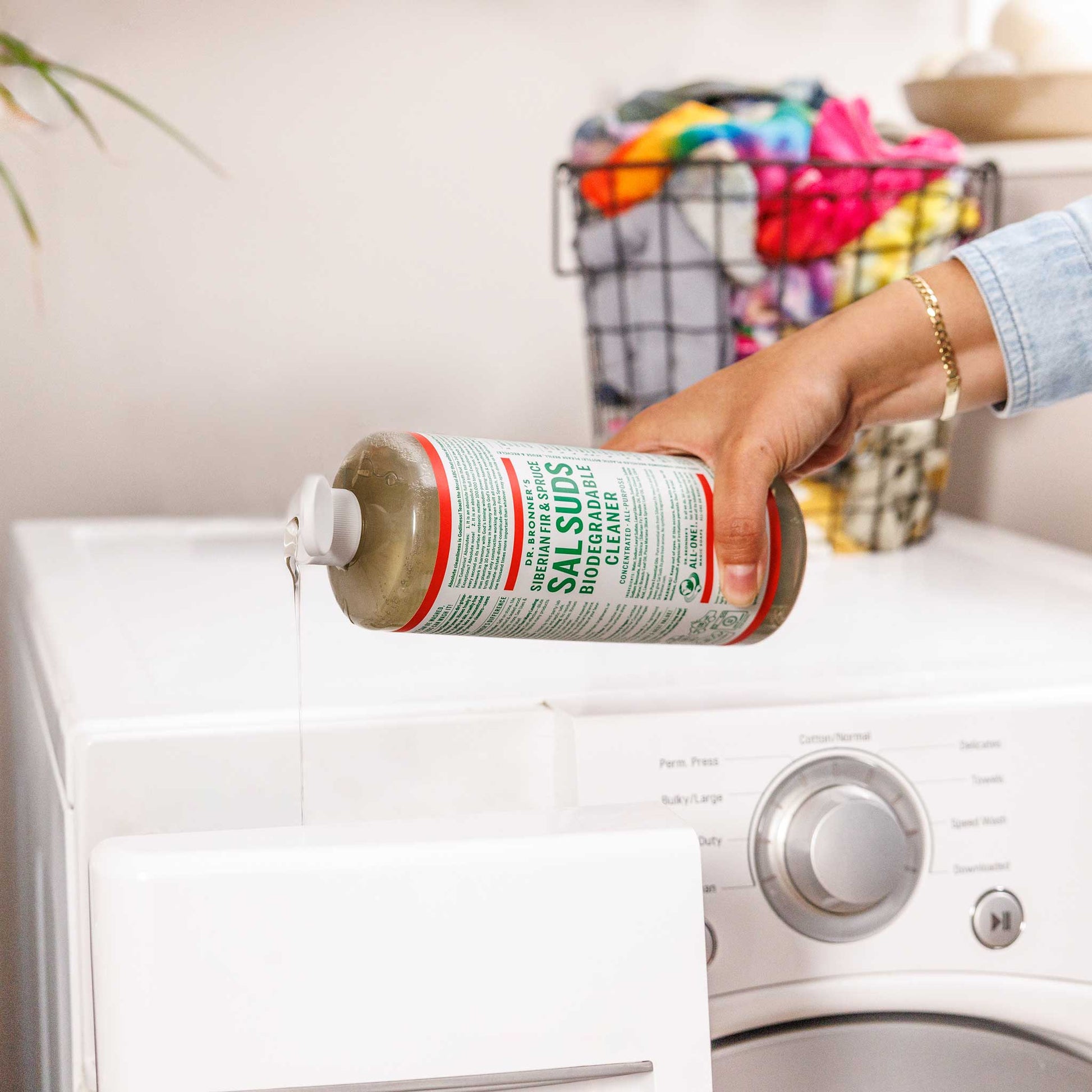 A hand pours Dr. Bronners liquid castile soap over a white laundry machine. Nearby, a basket filled with colorful clothes adds context to this natural cleaning scene [16 oz]
