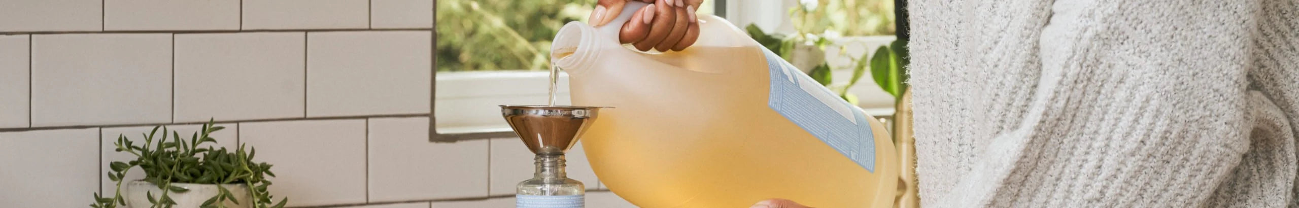 A person pours natural soap from a large container into a bottle using a funnel on a kitchen counter with white tiles and a potted plant nearby