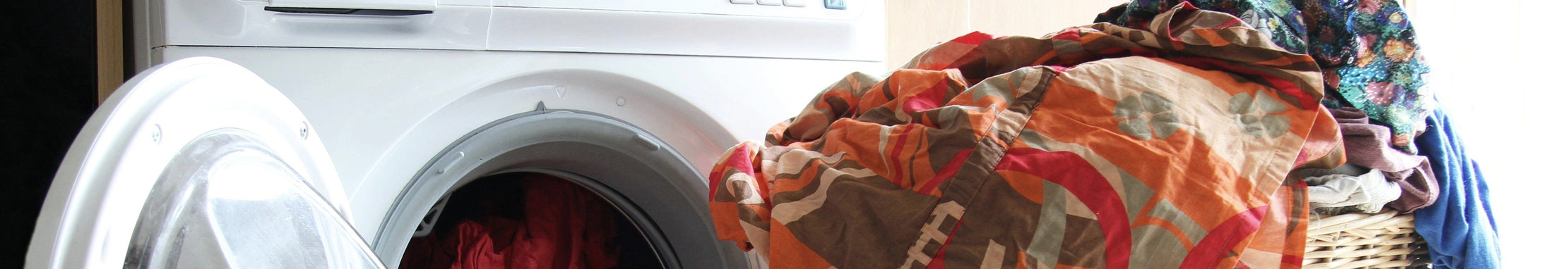 A front loading washing machine with the door open, revealing laundry inside. A colorful pile of clothes sits atop a wicker basket nearby, in a well lit laundry room
